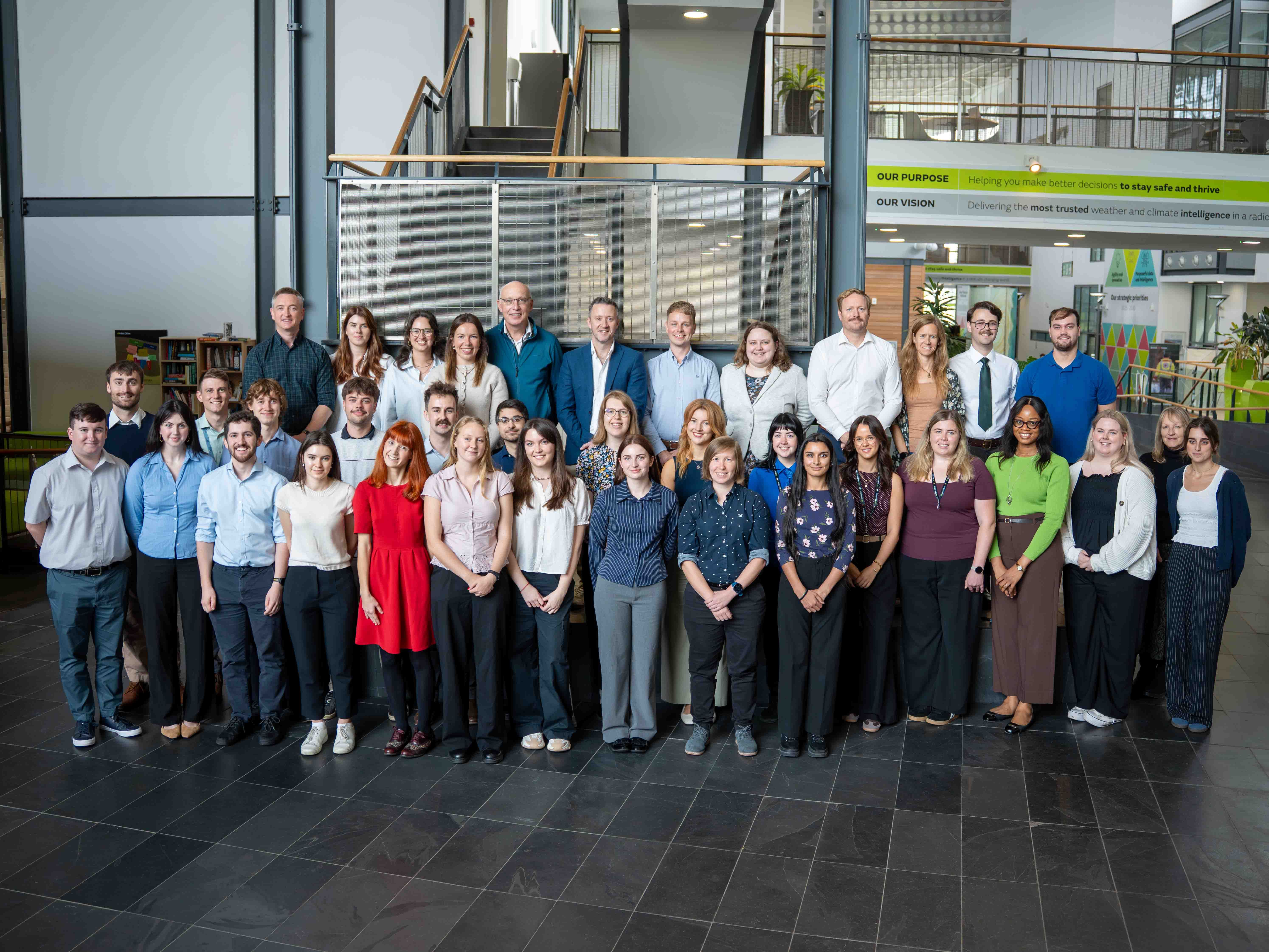 Large group photograph of the first cohort of the Foundation Operational Meteorology Programme standing in three rows inside the Met Office HQ in Exeter