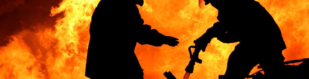 Silhouettes of two firefighters fighting a fire, one holding a hose spraying water on the ground, whilst a fire rages behind them
