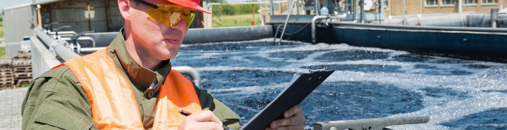 An engineer wearing a red hard hat and an orange hi-vis jacket writes on a clipboard while inspecting a water treatment plant.