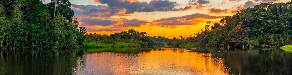 A view up a stretch of river towards a sunset on the horizon, in a tropical region with areas of dense forest on both sides.