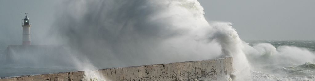 Storm waves crashing over harbour wall, with lighthouse in the distance looking small in comparison