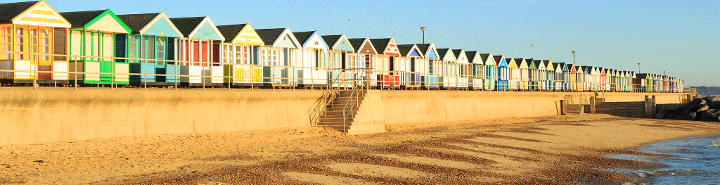 Beach huts on a promenade above sea defences, viewed from the shoreline with a pebble beach