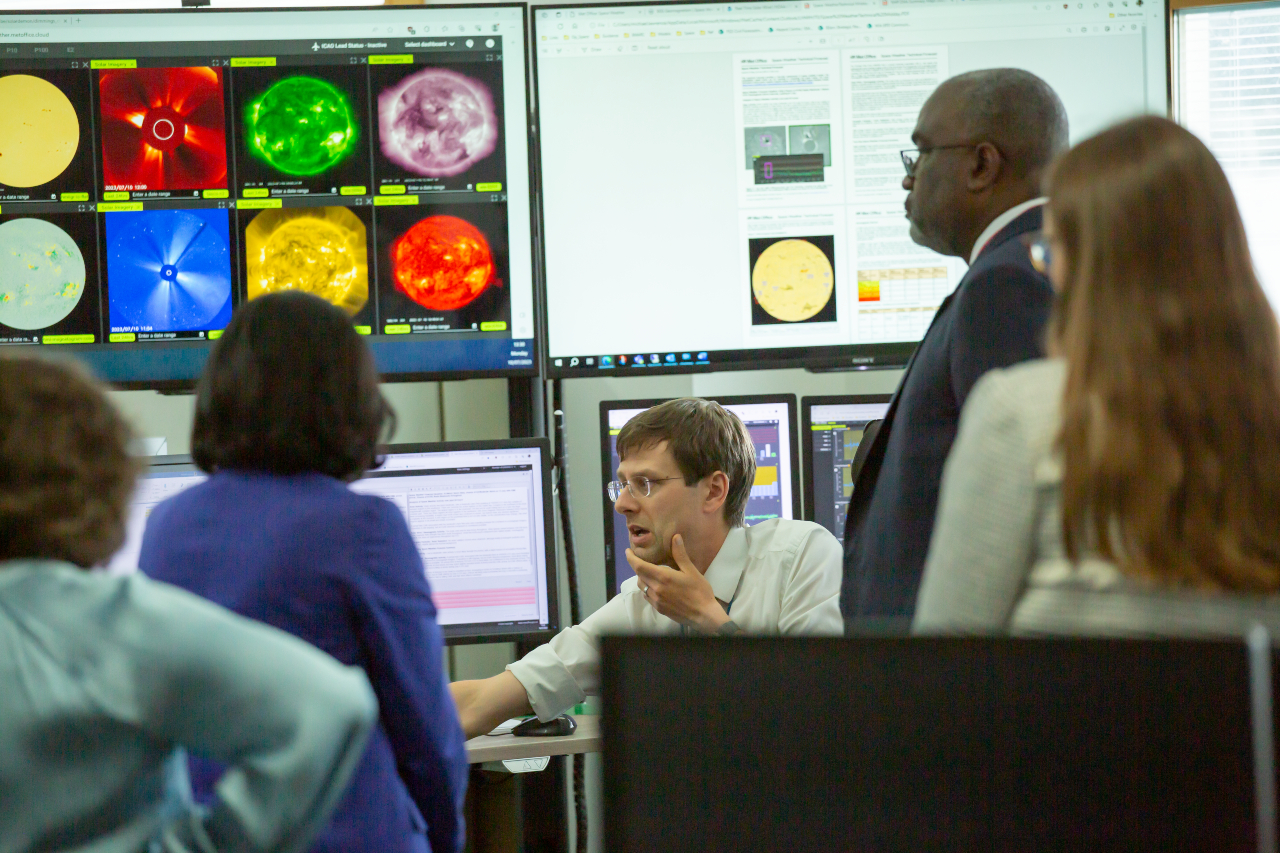 Space weather section in Met Office HQ showing a TV with various images of the sun, Dr Morgan being shown the images by Met Office staff.