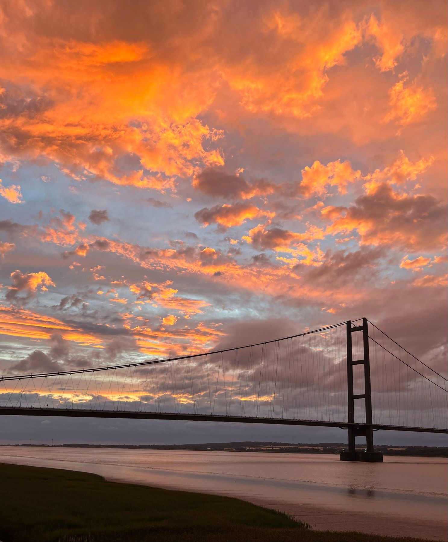 A photo from one bank of a river crossed by a suspension bridge at sunset, with skies coloured orange, pink, yellow, blue and grey