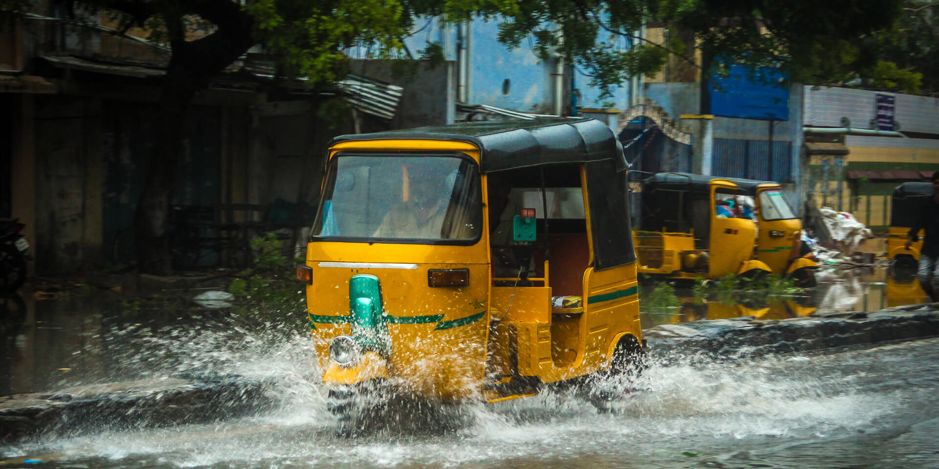 A yellow tuk tuk driving through flood water creating a splash
