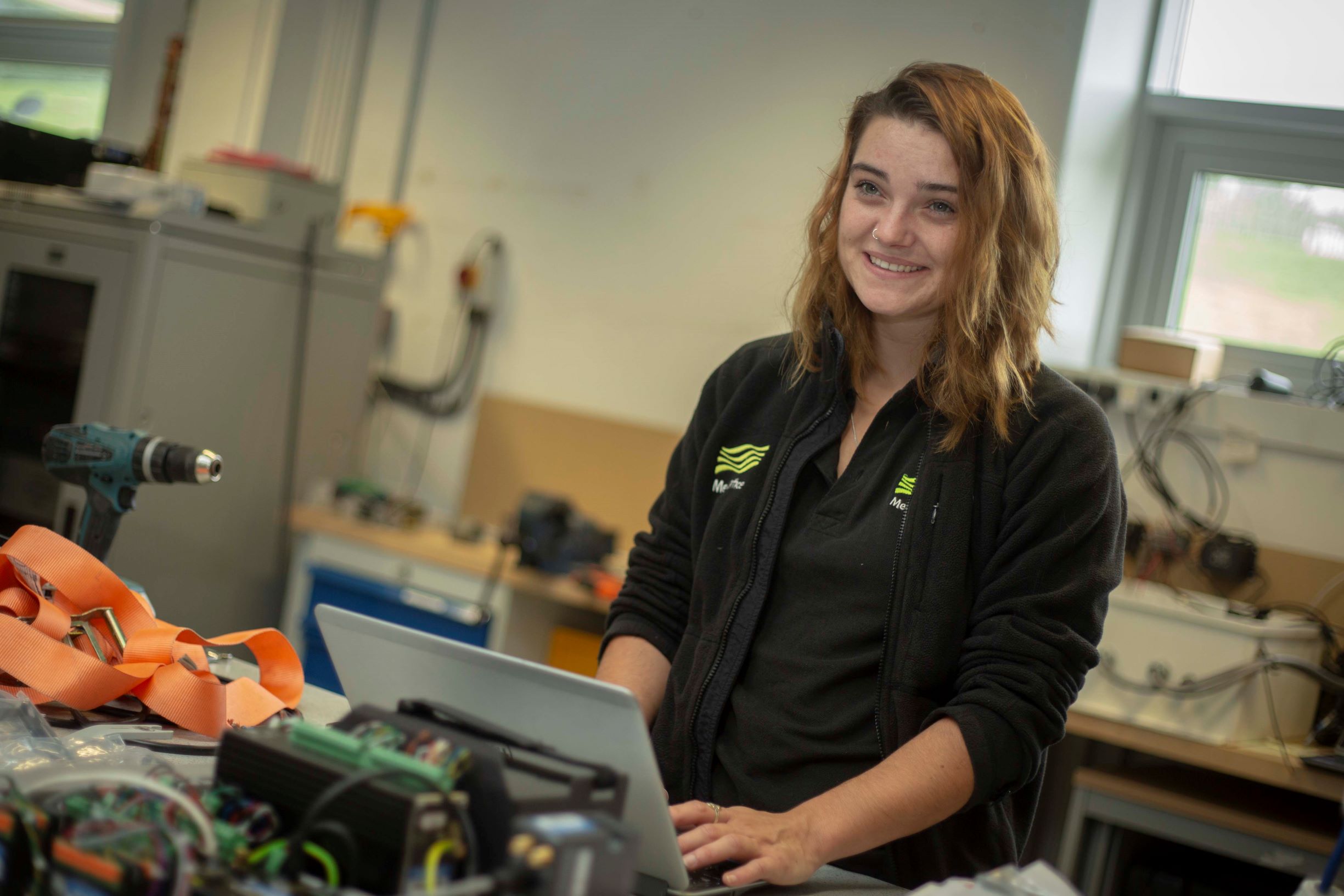 Tess Brock using her laptop in a workshop setting, surrounded by tools and equipment.